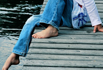 Barfüßiges Kind in Jeans sitzt auf einem Steg und streckt einen Fuß ins Wasser, Foto: kallejipp | Photocase.de
